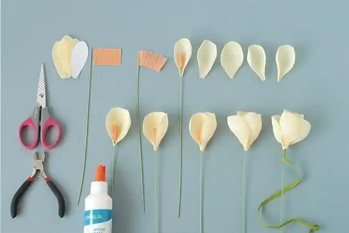 The image shows a step-by-step process for creating paper flowers. The materials and tools laid out include: A pair of pink-handled scissors with a patterned blade. A pair of black-handled pliers. A bottle of glue labeled "ORALIA." Several pieces of paper cut into petal shapes. Strips of orange paper with fringed edges. Green floral wires. Partially assembled paper flowers in various stages of completion.
