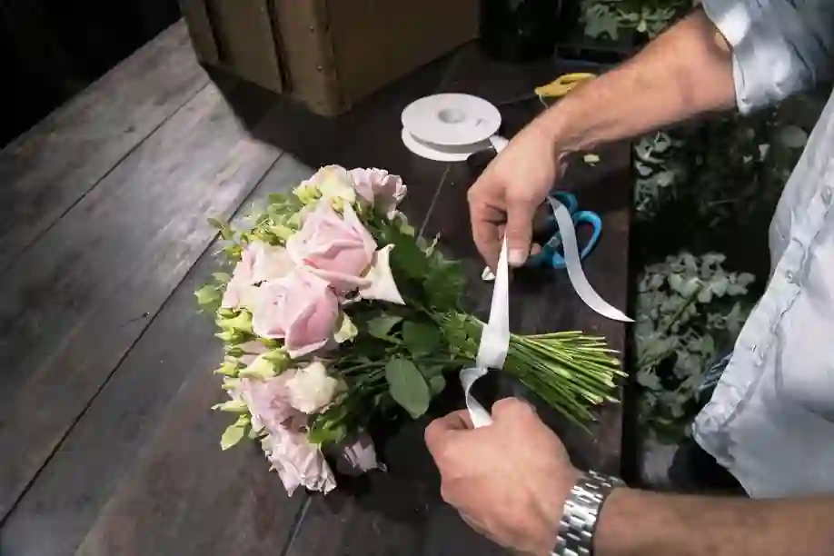 The image shows a person arranging a bouquet of flowers on a wooden table. The person is tying a white ribbon around the stems of the bouquet, which consists of pink and white flowers with green leaves. On the table, there are also a pair of blue-handled scissors, a roll of white ribbon, and some other tools. The person is wearing a light-colored shirt and a watch on the left wrist. The setting appears to be a workspace for flower arrangement or a florist's shop. This image is interesting as it captures the detailed process of creating a floral arrangement, highlighting the craftsmanship involved in preparing a bouquet.