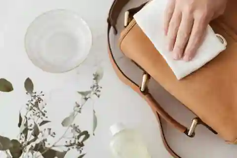 The image shows a hand cleaning a brown leather bag with a white cloth. The bag is placed on a white surface, and there is a clear bowl of water, a bottle of cleaning solution, and some green foliage nearby. The scene suggests a process of maintaining or cleaning the leather bag, which is interesting as it highlights the care and upkeep required for leather goods. Cleaning and maintaining leather items like this bag is essential to keep them looking great and lasting longer. Using the right products and techniques can help preserve the leather's natural beauty and prevent damage.