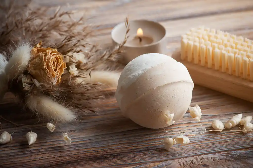 The image shows a collection of items arranged on a wooden surface. The items include a bath bomb, a dry brush, a lit candle in a small holder, and a bouquet of dried flowers. The bath bomb is spherical and light beige in color. The dry brush has a wooden handle and stiff bristles. The candle is lit, providing a warm glow, and is placed in a small, round holder. The bouquet of dried flowers includes various types of dried grasses and a large, dried rose. Small dried flower buds are scattered around the bath bomb. The overall setting suggests a relaxing, spa-like atmosphere.