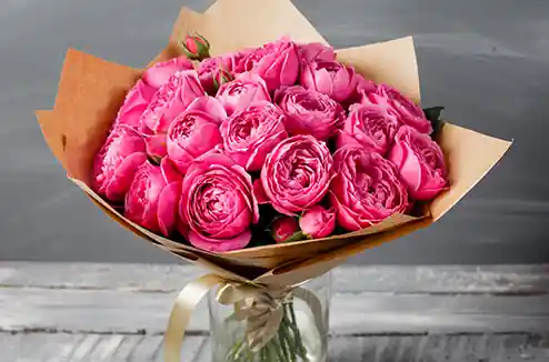 The image showcases a beautiful bouquet of pink roses, wrapped in brown paper and tied with a ribbon. The bouquet is placed in a glass jar, and the roses appear fresh with vibrant pink petals. The background is a grayish surface, possibly a wall, and the bouquet is resting on a wooden surface.