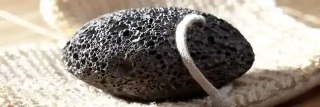 The image shows a black pumice stone with a white string looped through it, resting on a textured beige surface, possibly a washcloth or bath mat. The pumice stone has a porous texture, which is characteristic of volcanic rock. This type of stone is commonly used for exfoliating skin, particularly on the feet, to remove dead skin cells and calluses. The presence of the string suggests it can be hung up to dry after use, which is practical for maintaining hygiene. The image is interesting and relevant as it depicts a common personal care item used in skincare routines.