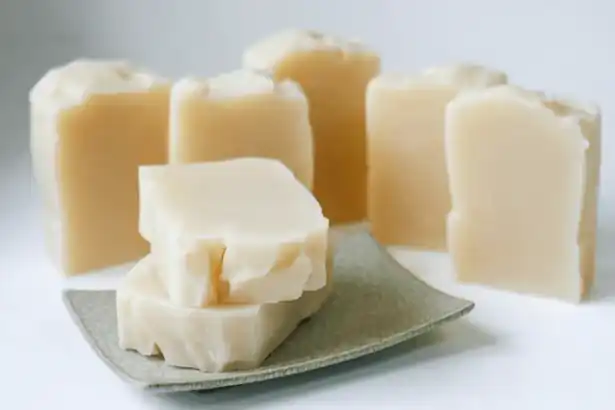 The image showcases several bars of soap arranged on a white surface. There are six bars in total, with two lying flat on a small, square dish in the foreground and four standing upright in the background. The soap bars have a light beige color and a slightly uneven, handmade appearance, indicating they are likely artisanal soaps. They look quite natural and appealing, suggesting they may be crafted with care and natural ingredients.
