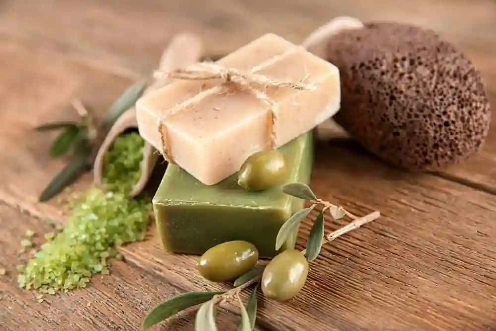 The image shows a rustic arrangement of natural bath products on a wooden surface. There are two bars of soap, one beige and one green, with the beige soap tied with a piece of twine. Surrounding the soaps are green olives, olive branches with leaves, a wooden spoon filled with green bath salts, and a pumice stone. The scene is set on a wooden table, giving it a natural and organic feel. This image is interesting and relevant as it highlights natural and eco-friendly bath products, which are popular for their health and environmental benefits.