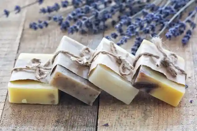 The image shows four bars of handmade soap, each wrapped in brown paper and tied with a piece of twine. The soaps are arranged in a staggered stack on a wooden surface. In the background, there are sprigs of dried lavender flowers. This image highlights natural, artisanal products that are often appreciated for their aesthetic appeal and potential benefits for skin care.
